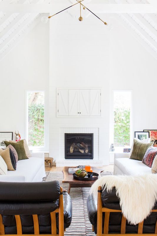 A modern farmhouse living room in white, with white sofas and black leather chairs, a fireplace and a TV behind white barn doors.
