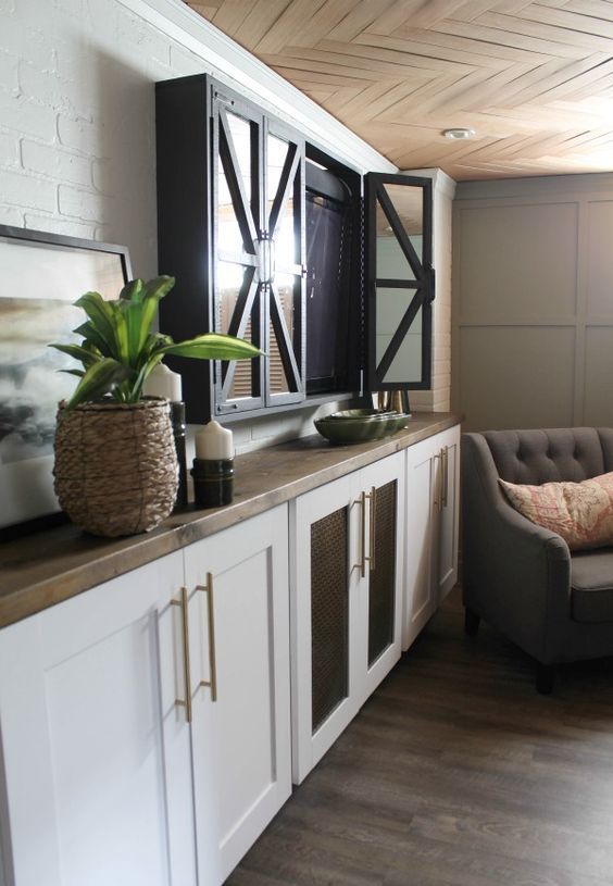 A modern living room with white cabinets, butcherblock countertops, a TV hidden behind doors with blinds and modern furniture.
