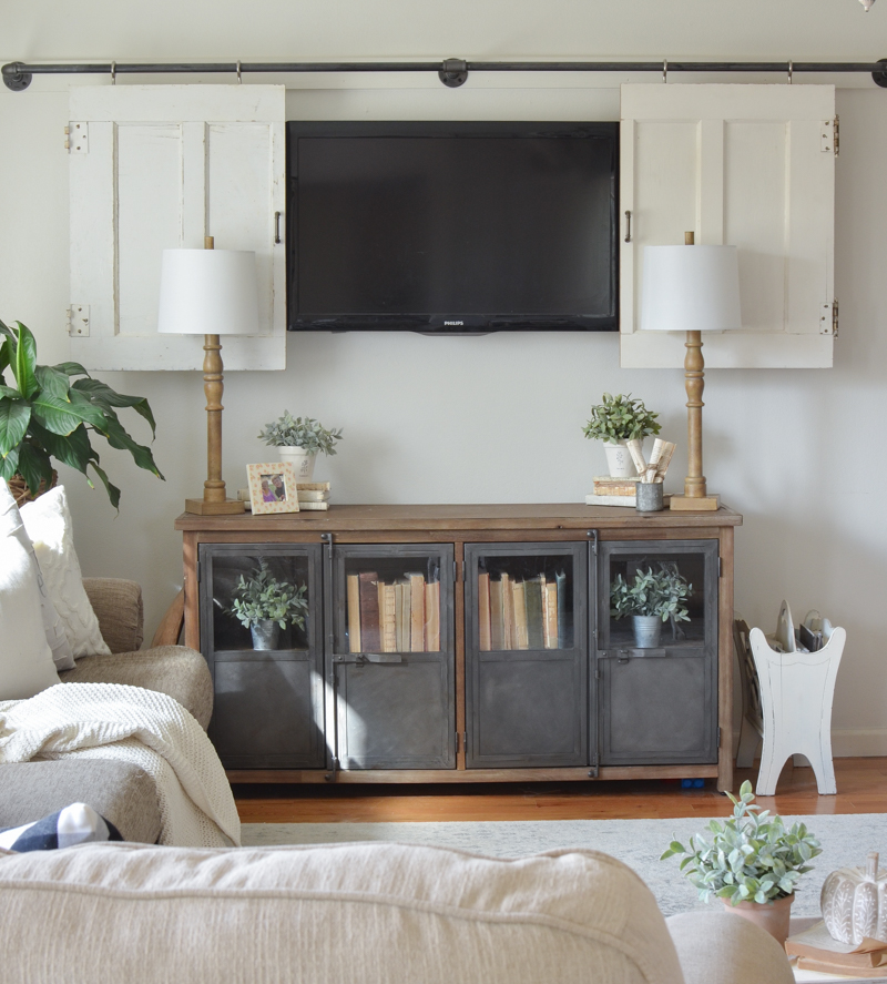 A neutral farmhouse living room with neutral sitting furniture, a TV on the wall hidden behind white barn doors and a metal and wood storage unit.