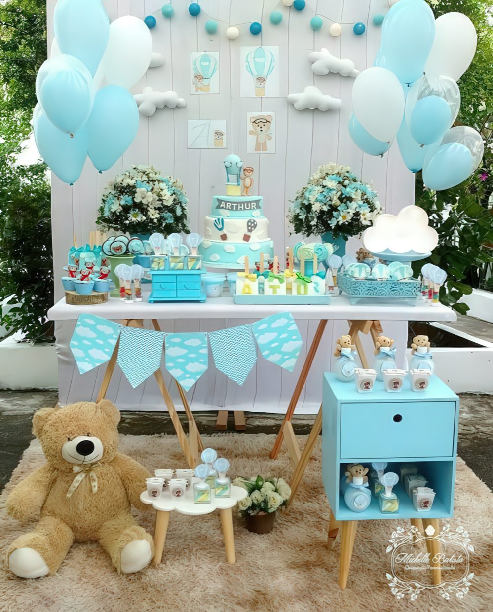 A blue dessert table with a backdrop done with clouds and blue balloons, a blue garland and blue and white floral arrangements.