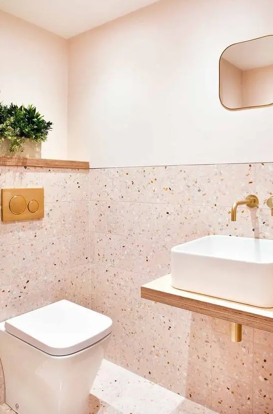 A floating vanity enhances the sleek look of this blush powder room with terrazzo tiles and paint. Gold fixtures and touches add sophistication. Choose floating furniture and terrazzo for a chic, contemporary design.