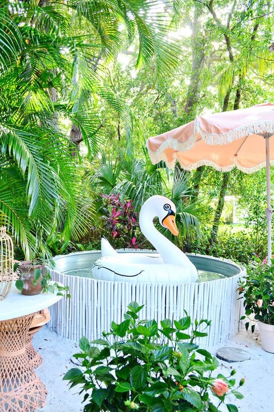 A bright and funny pool nook with a stock tank pool clad with a stick cover, potted greenery all around, a pink umbrella and rattan furniture.