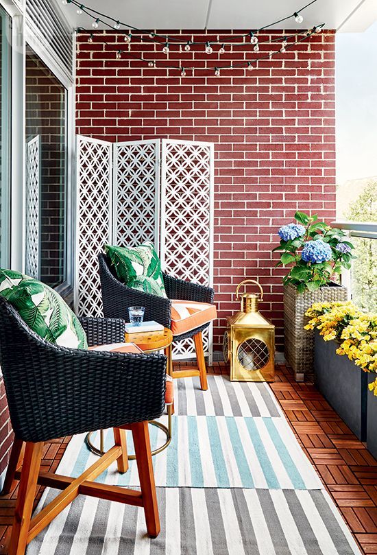 A bright and stylish balcony with layered rugs, wicker chairs and printed pillows, potted blooms and a large candle lantern.