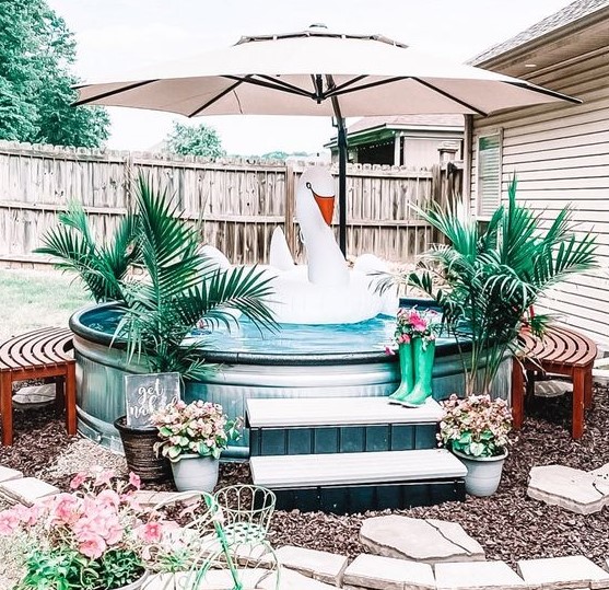 A brigth and cool pool space with a stock tank pool, wooden benches and a ladder, potted plants, an umbrella and a large float.