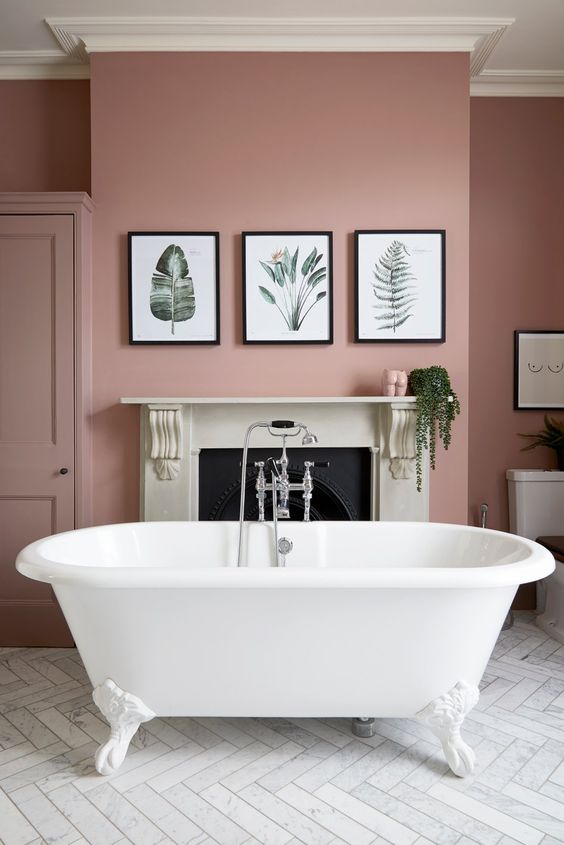 A chic pink bathroom with a non-working fireplace, a white clawfoot tub, a gallery wlal and some greenery.