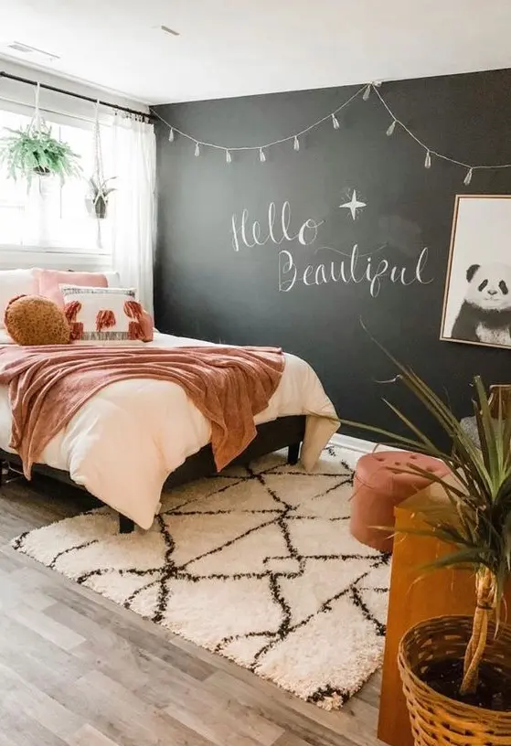 A chic teen bedroom with a chalkboard wall, a bed with pretty pink bedding, a wooden desk, a chair and a footrest.