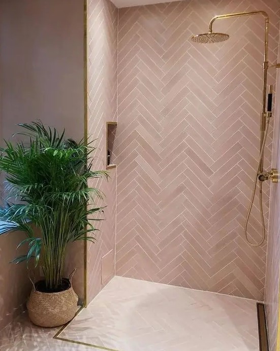 A contemporary pink bathroom clad with pink tiles in a herringbone pattern, gold fixtures and frames for a glam look.
