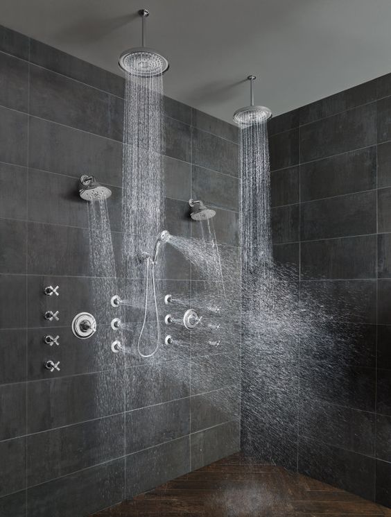 A contemporary shower space with large scale grey stone tiles, a dark stone chevron floor, rain shower heads on the ceiling and some on the wall.