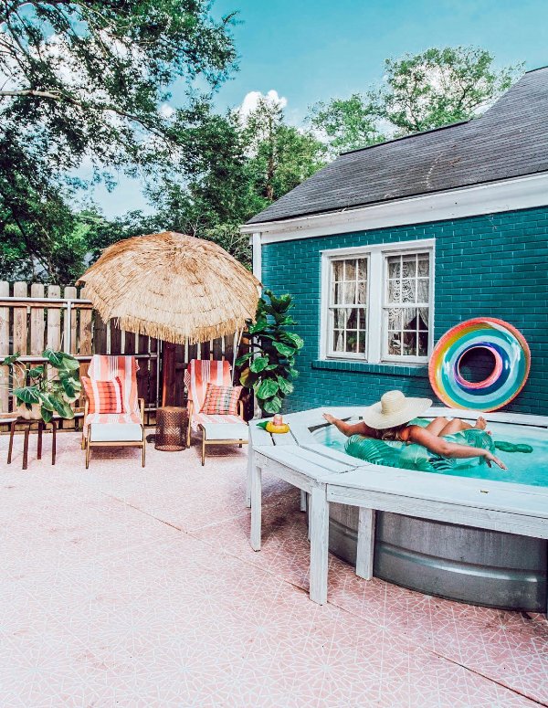 A cool and bright outdoor space with pink large format tiles, a stock tank pool with a wooden deck around it, loungers, an umbrella and bright floats.