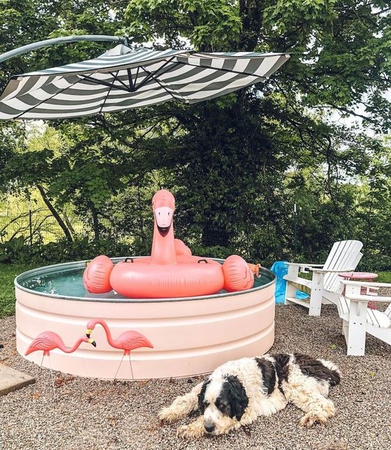 A cool pool space with a stock tank pool in blush, with a flamingo float, flamingos and a striped umbrella and outdoor chairs.