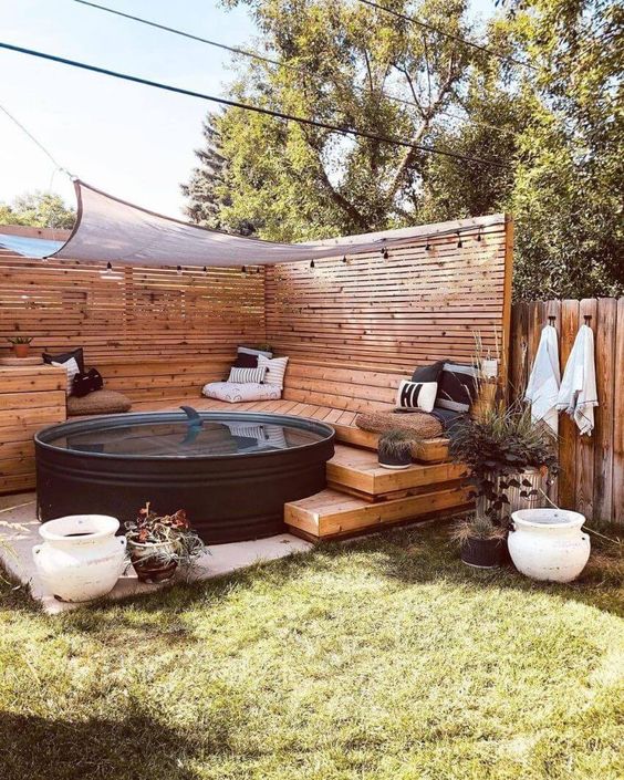 A cozy outdoor space with a stepped wooden deck with pillows, a black stock tank pool, a cover and lights.