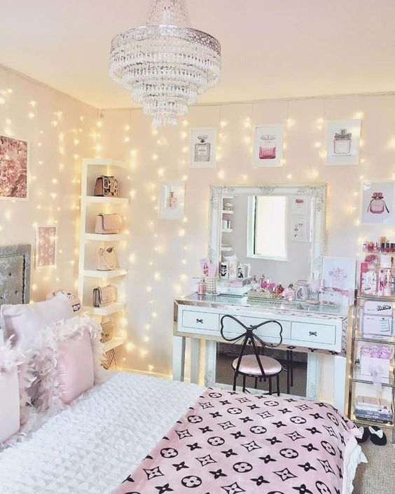 A glam teen bedroom with a grey bed with pink and blue bedding, a white desk and a mirror, a crystal chandelier and a shelf in the corner.