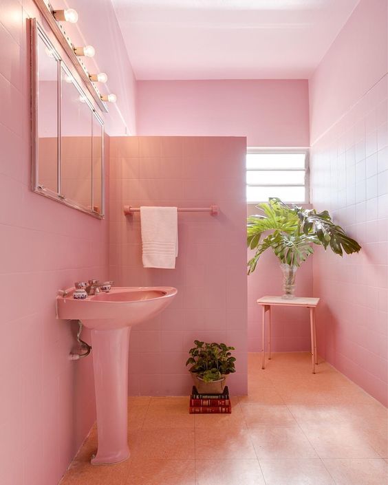 A light pink bathroom clad with square tiles, a free-standing sink, a shower space, a mirror with lights and potted greenery.