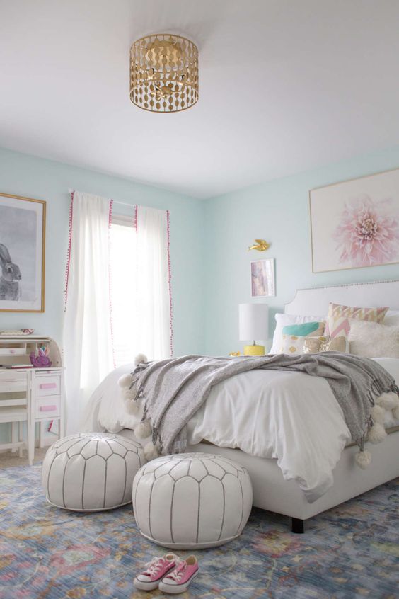 A lively teen girl bedroom with aqua walls, a white bed with printed pillows, white poufs, a white desk and a chair, a gold chandelier.