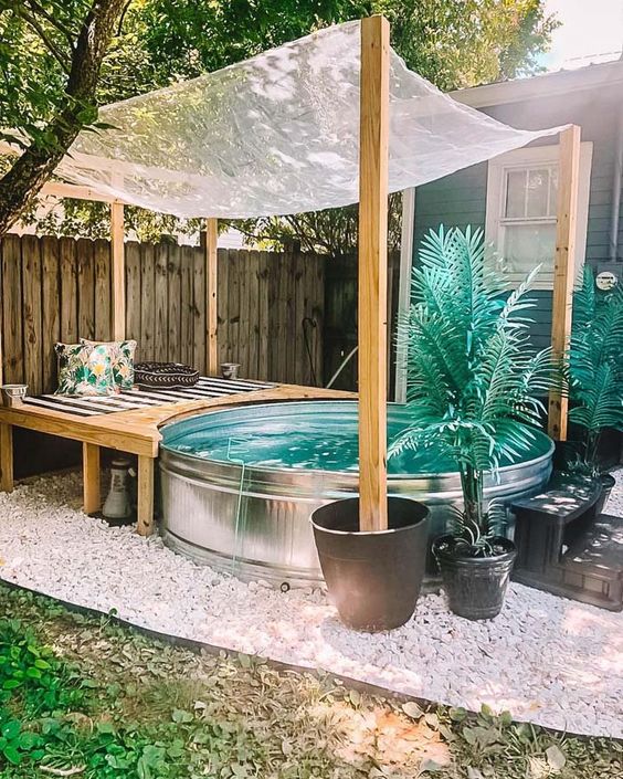 A lovely boho nook with a stock tank pool, a wooden bench with pillows, potted plants and gravel around.