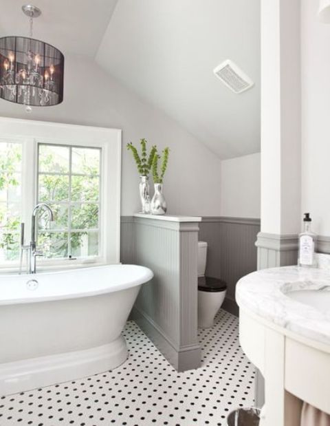 A lovely farmhouse bathroom with a tile floor, grey paneling on the walls, a vintage tub and a chic chandelier plus a half wall that separates the space.
