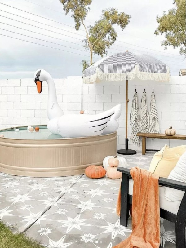 A lovely outdoor space with a tiled deck, a stock tank pool, wooden chairs and benches, an umbrella and some towels.