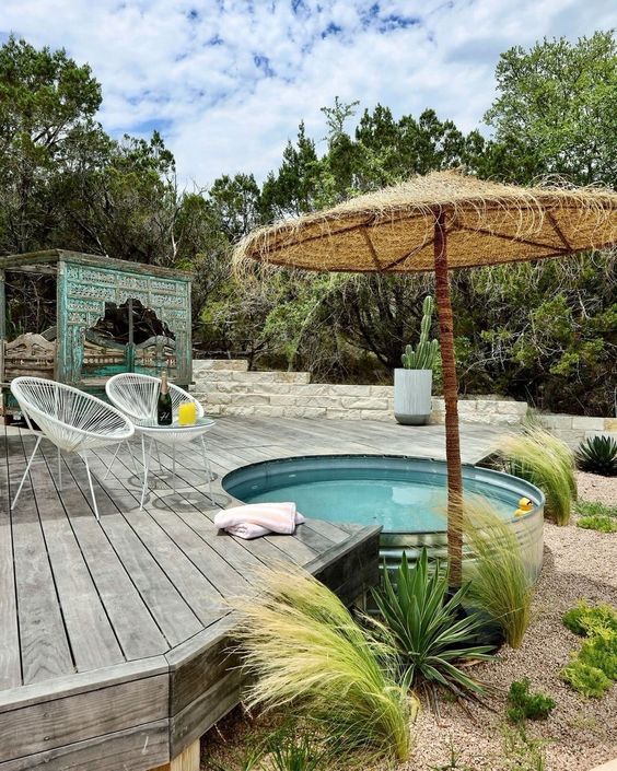 A lovely outdoor space with a wooden deck, a stock tank pool, white chairs, an umbrella and some greenery.