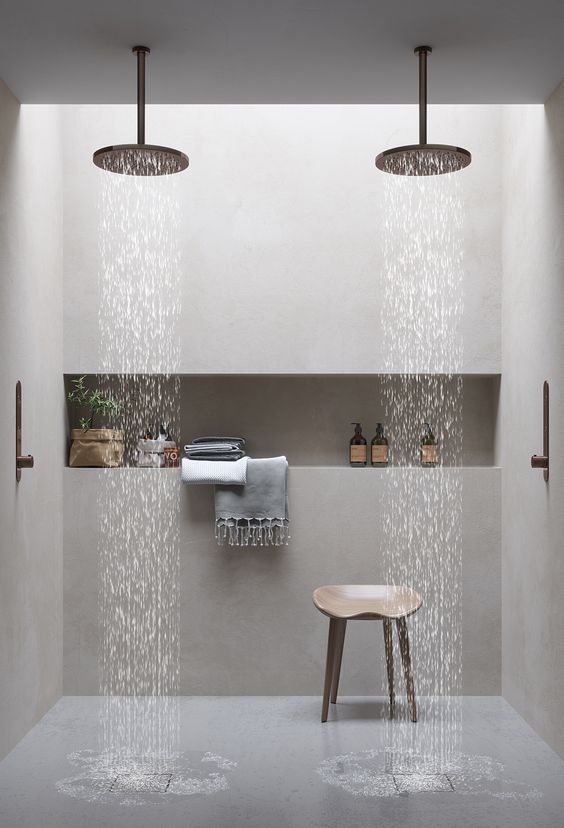 A minimalist bathroom done with neutral stone, with a niche shelf for storage, rain shower heads and a wooden stool.