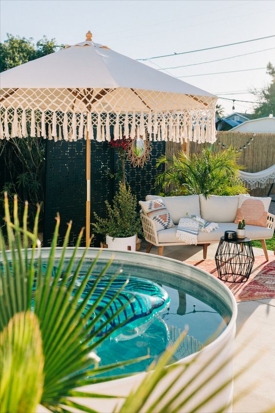 A modern boho space with a tiled deck, a stock tank pool, an umbrella with tassels, a sofa with pillows.
