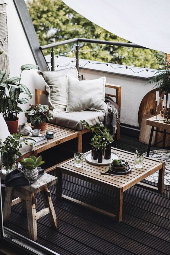 A monochromatic balcony with a black floor, natural wooden furniture, neutral textiles, potted greenery and candles.
