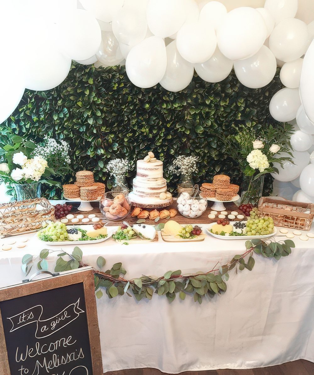 A neutral baby shower dessert table with a greenery backdrop, a large white balloon garland, a greenery garland and white blooms.