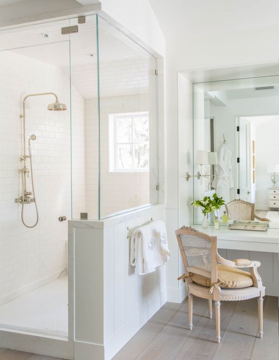 A neutral vintage bathroom with a shower space with a half wall, a vanity with a mirror and a vintage chair.