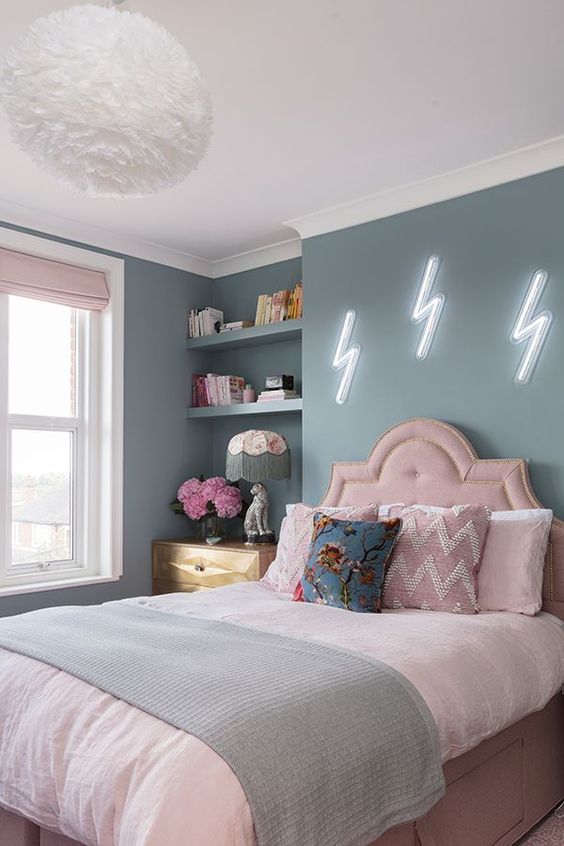 A pastel teen bedroom with blue walls, a pink bed with pastel bedding, built-in shelves, a stained dresser and blush curtains.