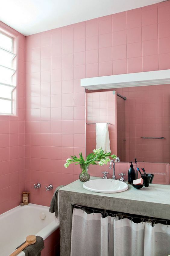 A pink bathroom clad with square tiles, a concrete vanity for a contrast, a mirror with a lamp and a bathtub is lovely.
