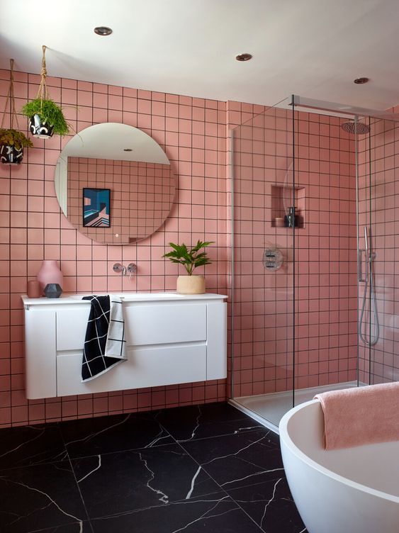 A pink bathroom with a black marble floor, white appliances and a floating vanity, potted greenery and a round mirror.