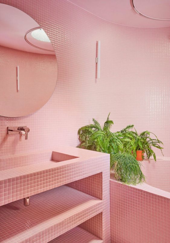 A pink bathroom with small scale tiles all over the space, a round mirror, potted greenery and a vanity clad with tiles, too.