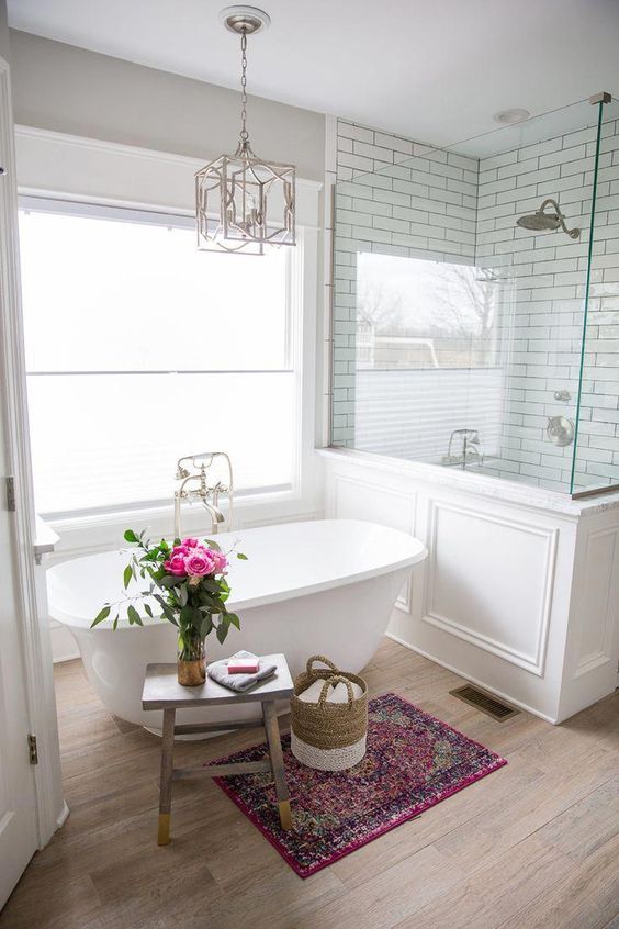 A refined farmhouse bathroom with a shower space with half walls, a small tub, a wooden stool and a chic chandelier.