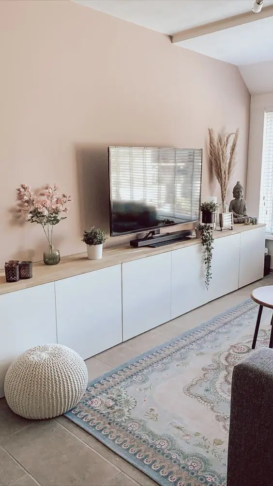 A Scandinavian living room with a blush wall, a large TV unit of IKEA Metod, with a wooden countertop, some decor and potted plants.