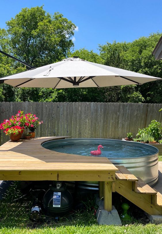 A simple outdoor space with a stock tank pool, a wooden deck with steps, an umbrella and some potted plants and blooms.