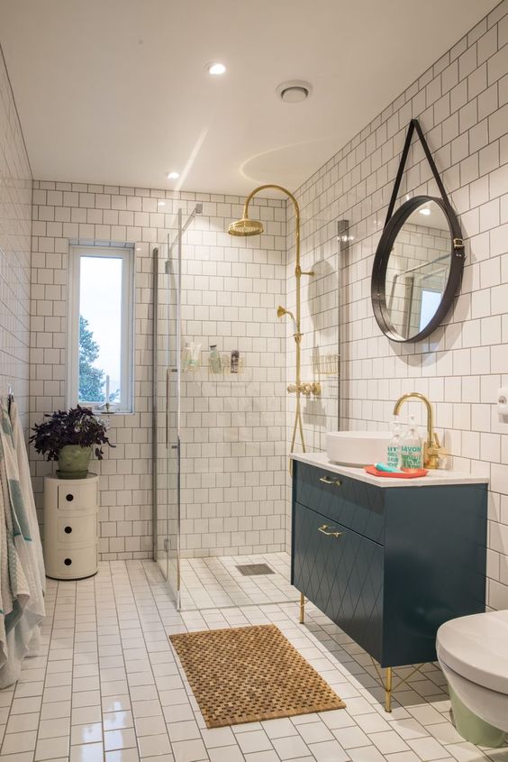A small and bright bathroom with white tiles, a navy vanity, a side table with a plant and touches of gold here and there.