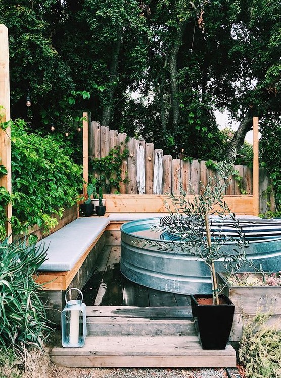 A small and cool space with two upholstered benches, a wooden deck, a stock tank pool with floats and potted plants.