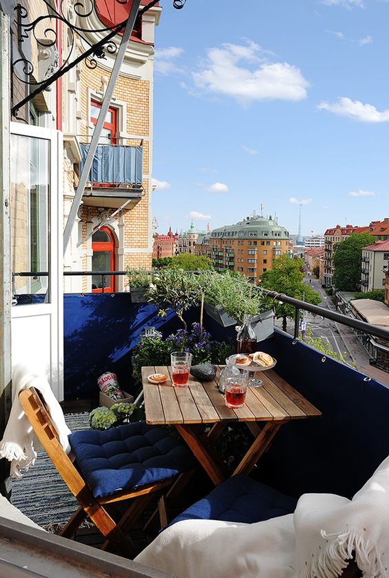 A small balcony with navy railing and upholstery, potted greenery and blooms and folding wooden furniture.
