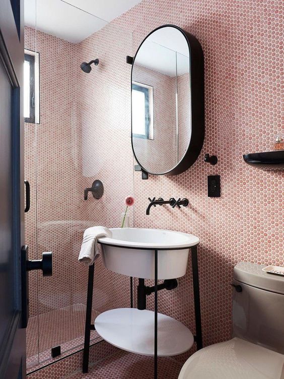 A small blush bathroom clad with pink penny tiles, a shower space, a creative sink on a stand, an oval mirror and black fixtures.