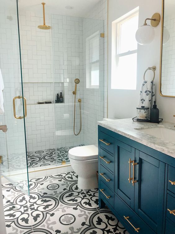 A small bright bathroom features white shower tiles and printed floor tiles. A navy vanity and brass fixtures complete the vibrant design.