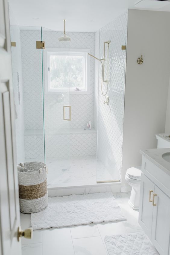 A small white bathroom features large scale and fish scale tiles. The shower space includes a white vanity, a basket, and gold fixtures.
