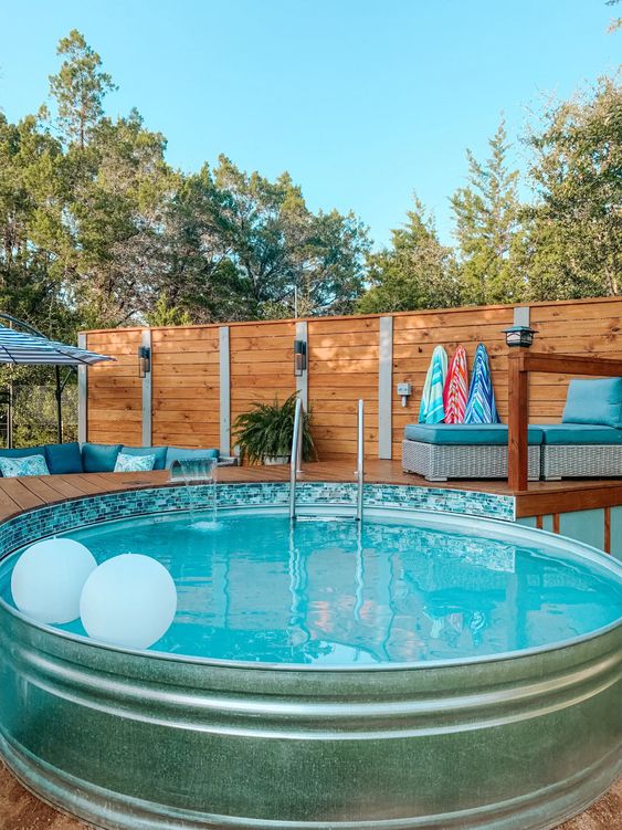A stock tank pool with floats, a wooden deck with a daybed, some greenery and bright towels.