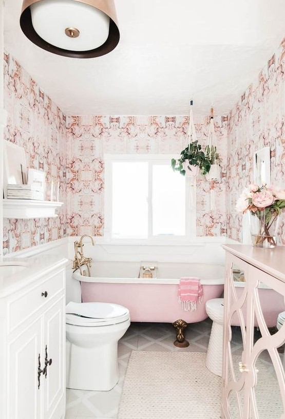 A stylish vintage-inspired bathroom done with pink printed wallpaper, a pink bathtub, a pink sideboard and brass fixtures and legs.