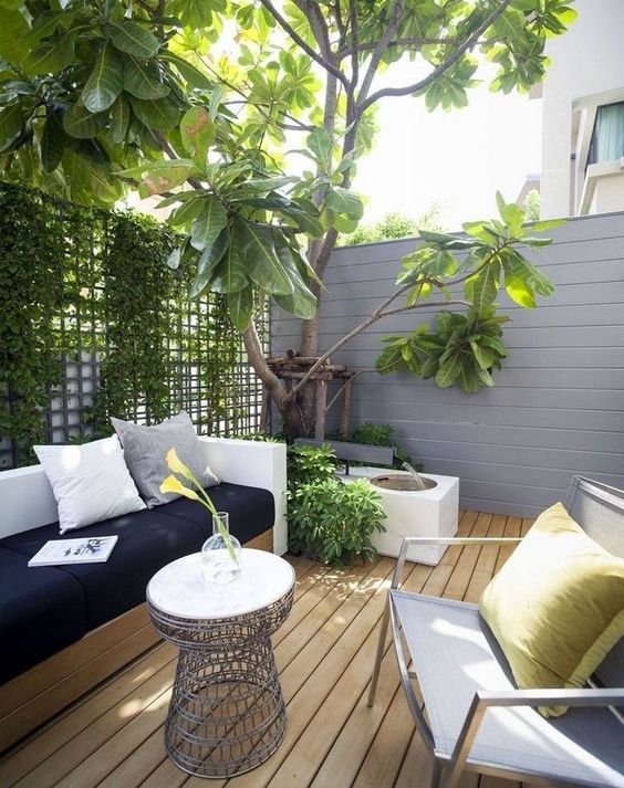 A tiny modern terrace with a wooden deck, a wall with greenery, a built-in sofa, a chair and a fountain plus a small table.