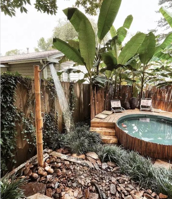 A tropical outdoor space with a lot of greenery and rocks around, a stock tank pool clad with bamboo, with a wooden deck and a bamboo shower.