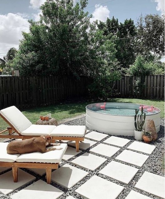 A welcoming backyard with gravel and tiles, loungers, candle lanterns, a white stock tank pool with floats and potted plants.