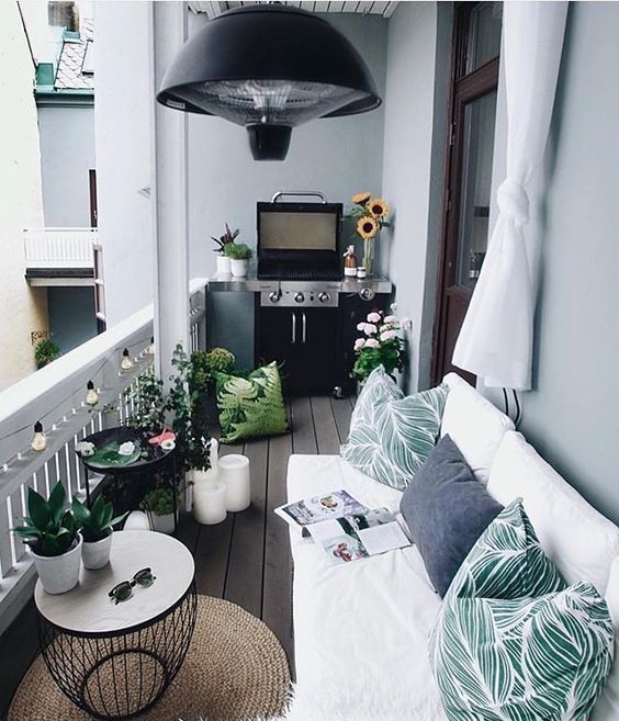 A welcoming balcony with a white L-shaped sofa, printed pillows, a grill, potted greenery and blooms.