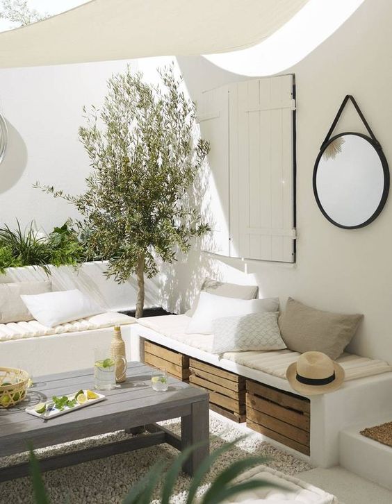 A white Mediterranean terrace with built-in benches, greenery and a tree, crates for storage and a small roof.