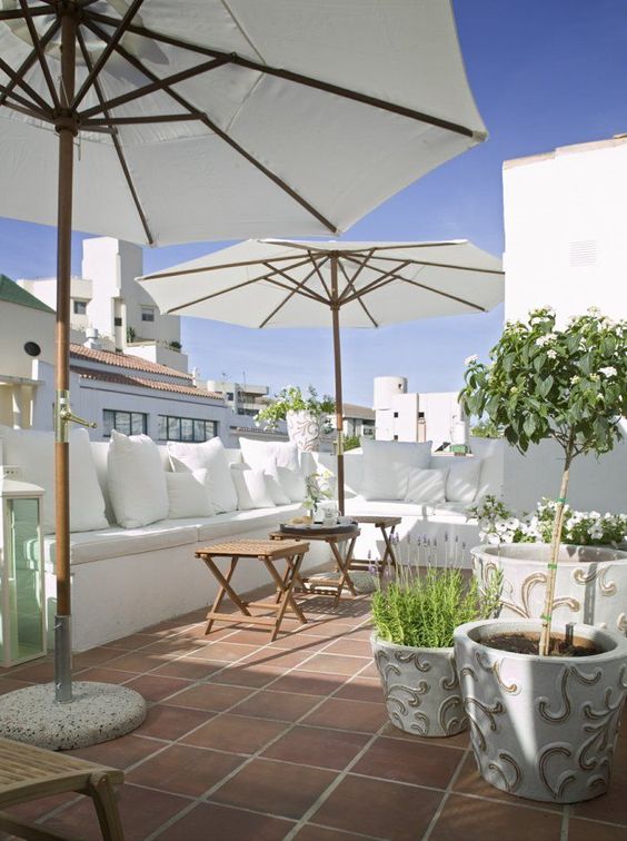 A white summer terrace with a built-in bench, white pillows, pottted blooms and greenery and umbrellas.