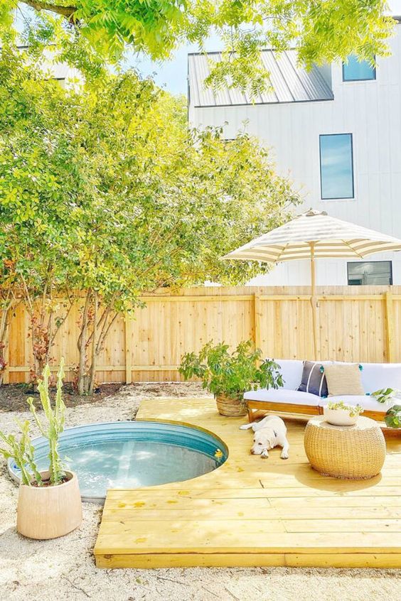 A wooden deck, an in-ground stock tank pool, a white sofa with pillows, potted plants and a pouf.