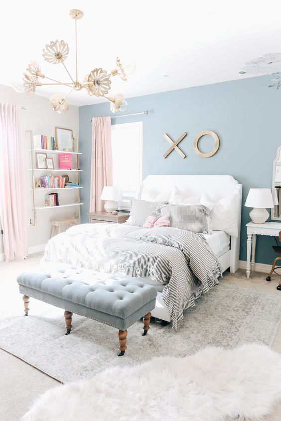 An airy teen girl bedroom with an ocean blue accent wall, open shelves in the corner, a white bed with neutral bedding, a powder blue upholstered bench.